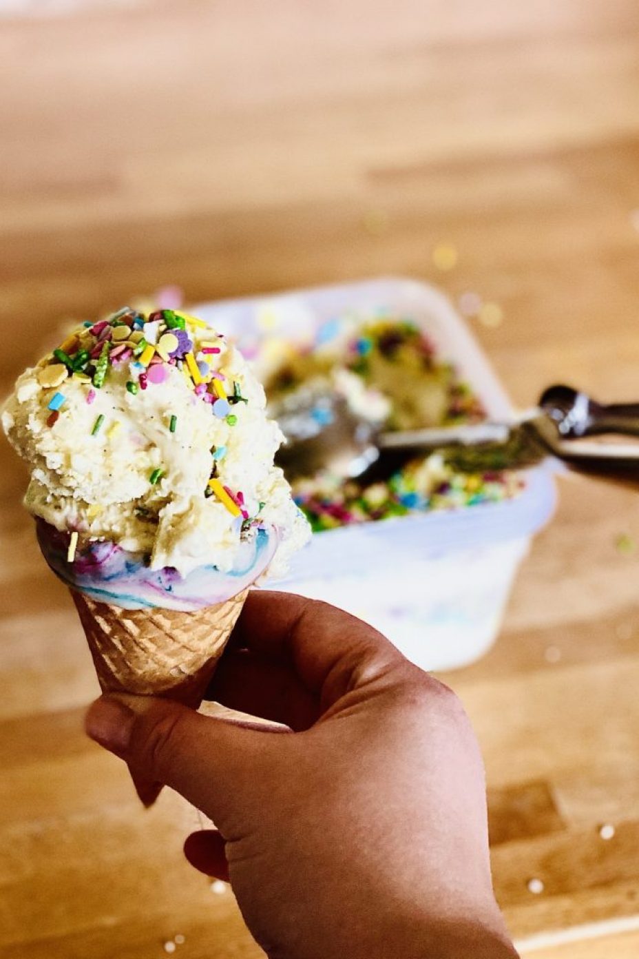 A scoop of white chocolate sparkle ice cream in an ice cream cone decorated with melted white chocolate. The ice cream tub with remaining ice cream behind.