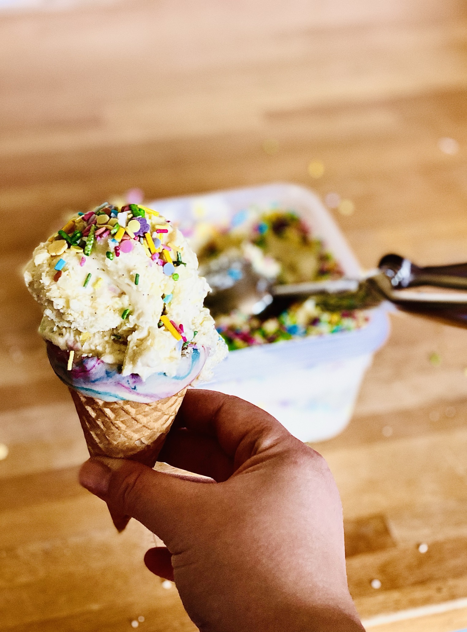 A scoop of white chocolate sparkle ice cream in an ice cream cone decorated with melted white chocolate. The ice cream tub with remaining ice cream behind.