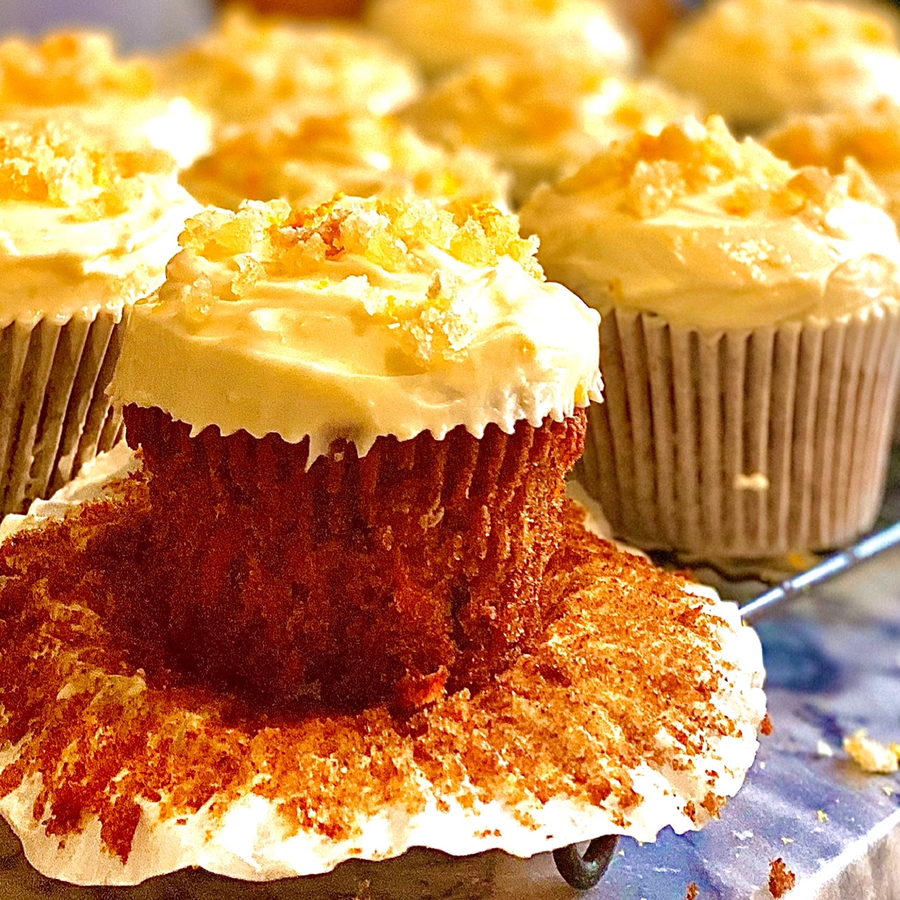 Lemon and ginger carrot cake cupcakes on a baking wrack. The front cake as the cake case turned down to show the carrot cake below contrasted with the pale cream cheese frosting.