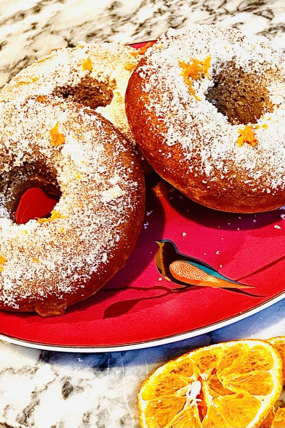 Baked banana and cinnamon breakfast doughnuts with orange sugar. Displayed on a plate with orange slices below for decoration.